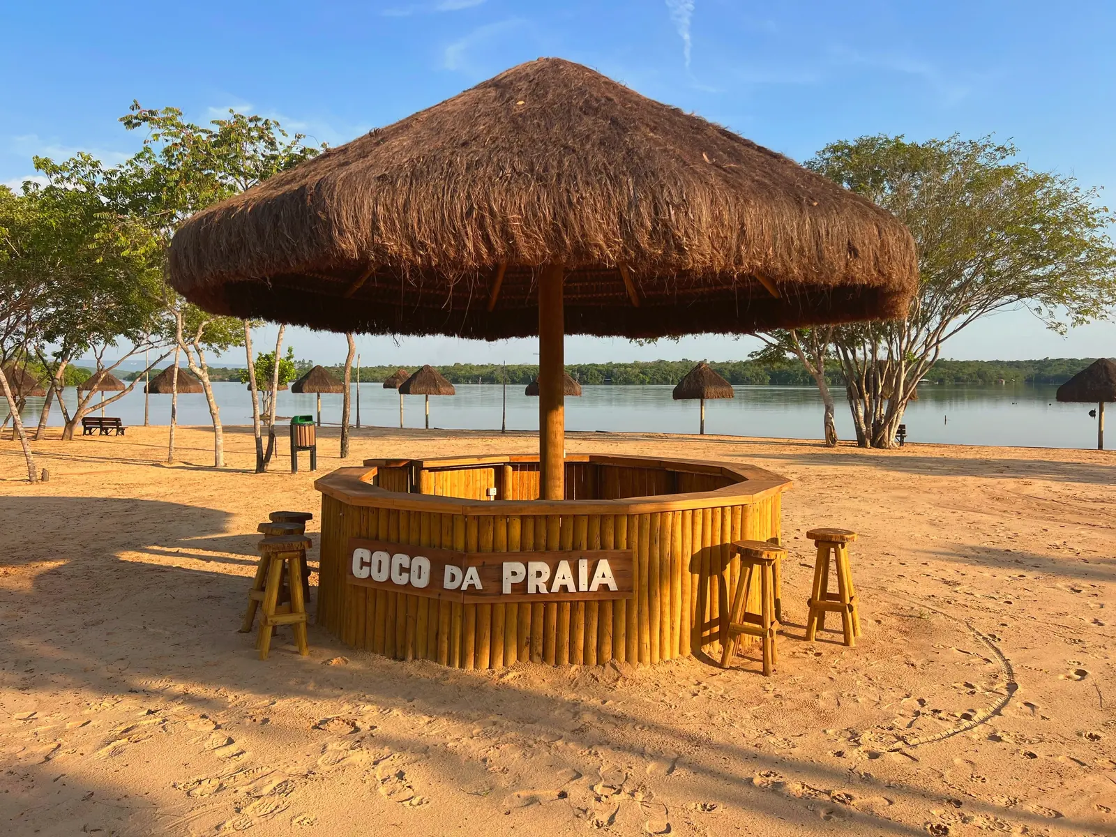Bar de praia coberto de palha na areia do Quinta do Lago, com bancos altos e o lago ao fundo